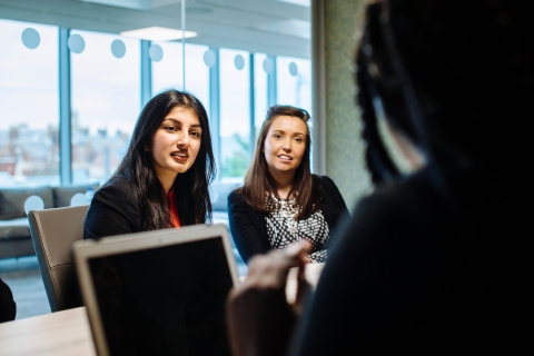 Two women sat at desk talking to woman in front of laptop